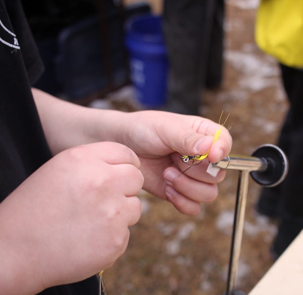 A fly tying vise holding a hook, with thread bobbin and materials on the bench